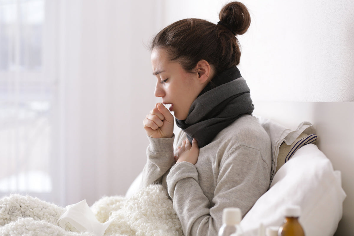 Eine junge Frau sitzt auf einem Bett mit einem Schal um ihren Hals. Sie hustet in ihre Hand und scheint Bronchitis zu haben.